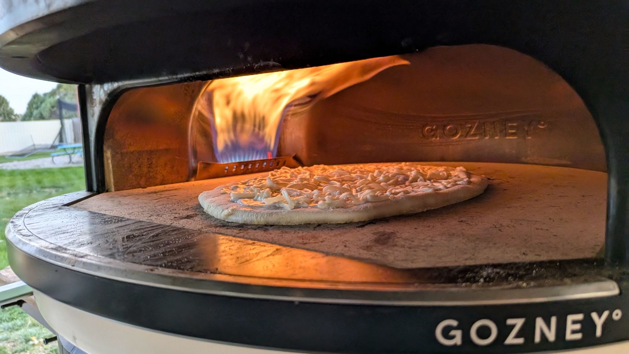 A cheese pizza baking inside the Gozney Arc XL with a large rolling gas flame on the left side.