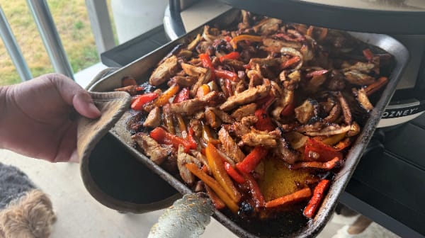 Removing a large sheet pan of sizzling chicken fajitas from the Gozney Arc XL pizza oven using a protective glove.