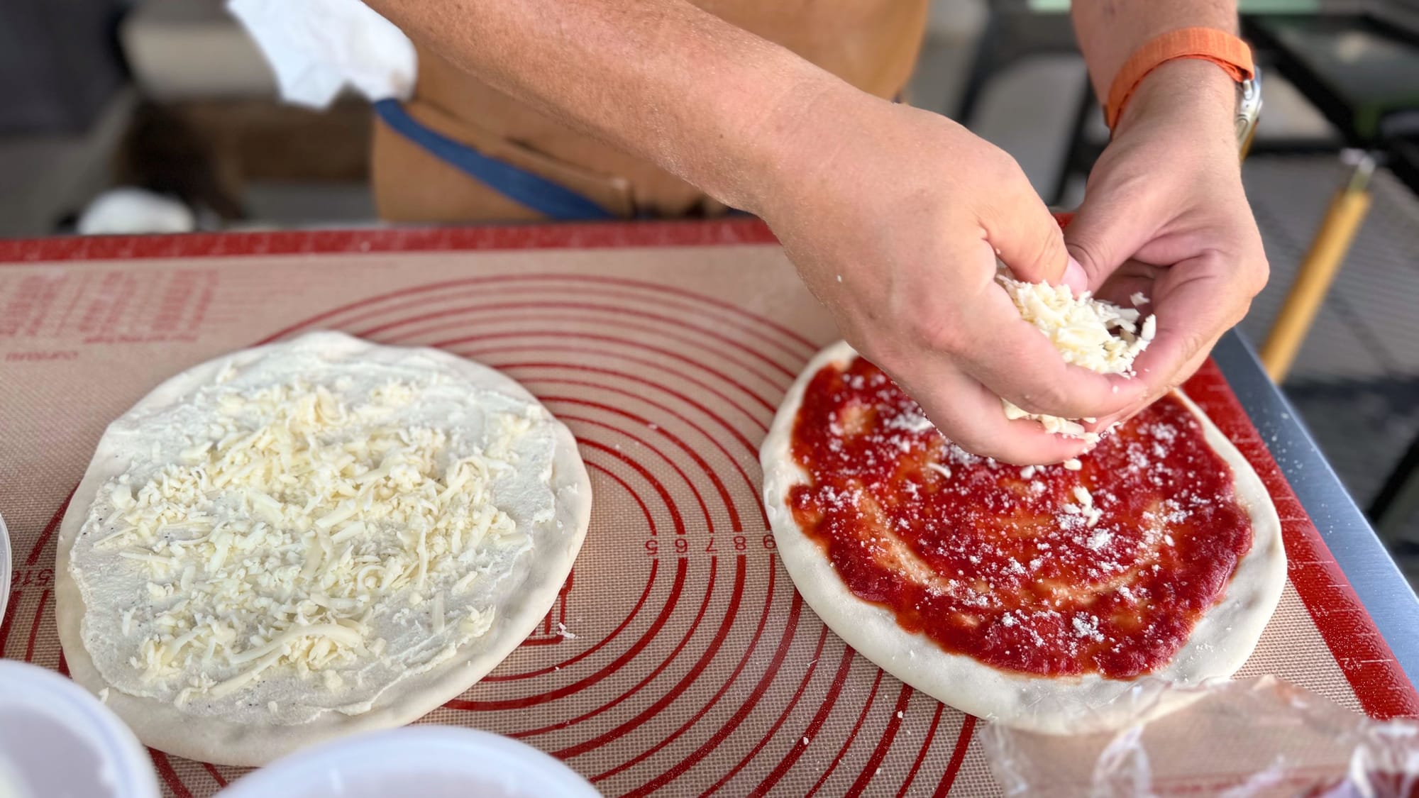 Preparing two pizzas on a silicone prep mat, sprinkling mozzarella cheese onto a fresh dough base with red sauce.