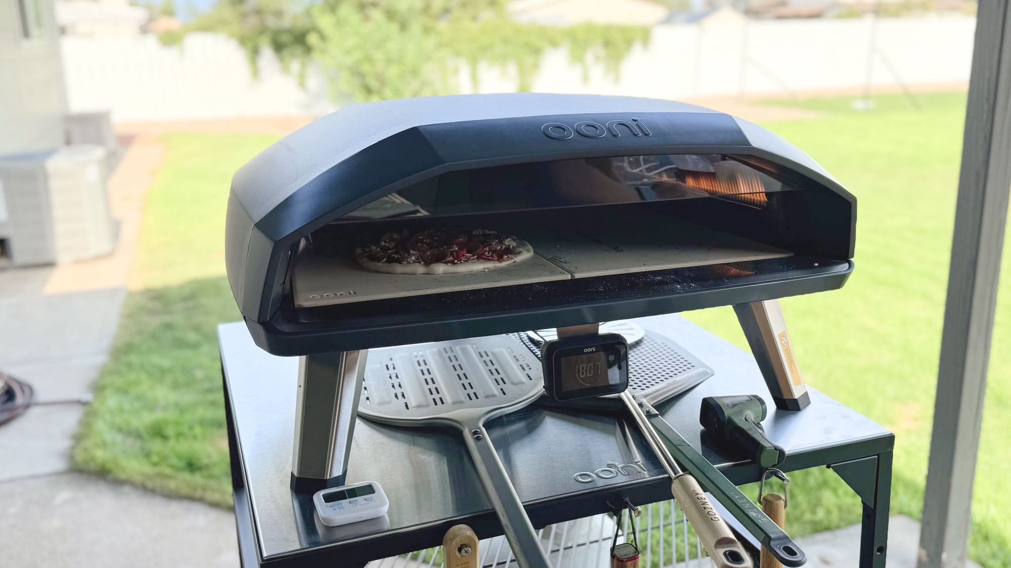 A pepperoni pizza baking inside the Ooni Koda 2 Max gas pizza oven, set up on an outdoor metal prep table with pizza peels.