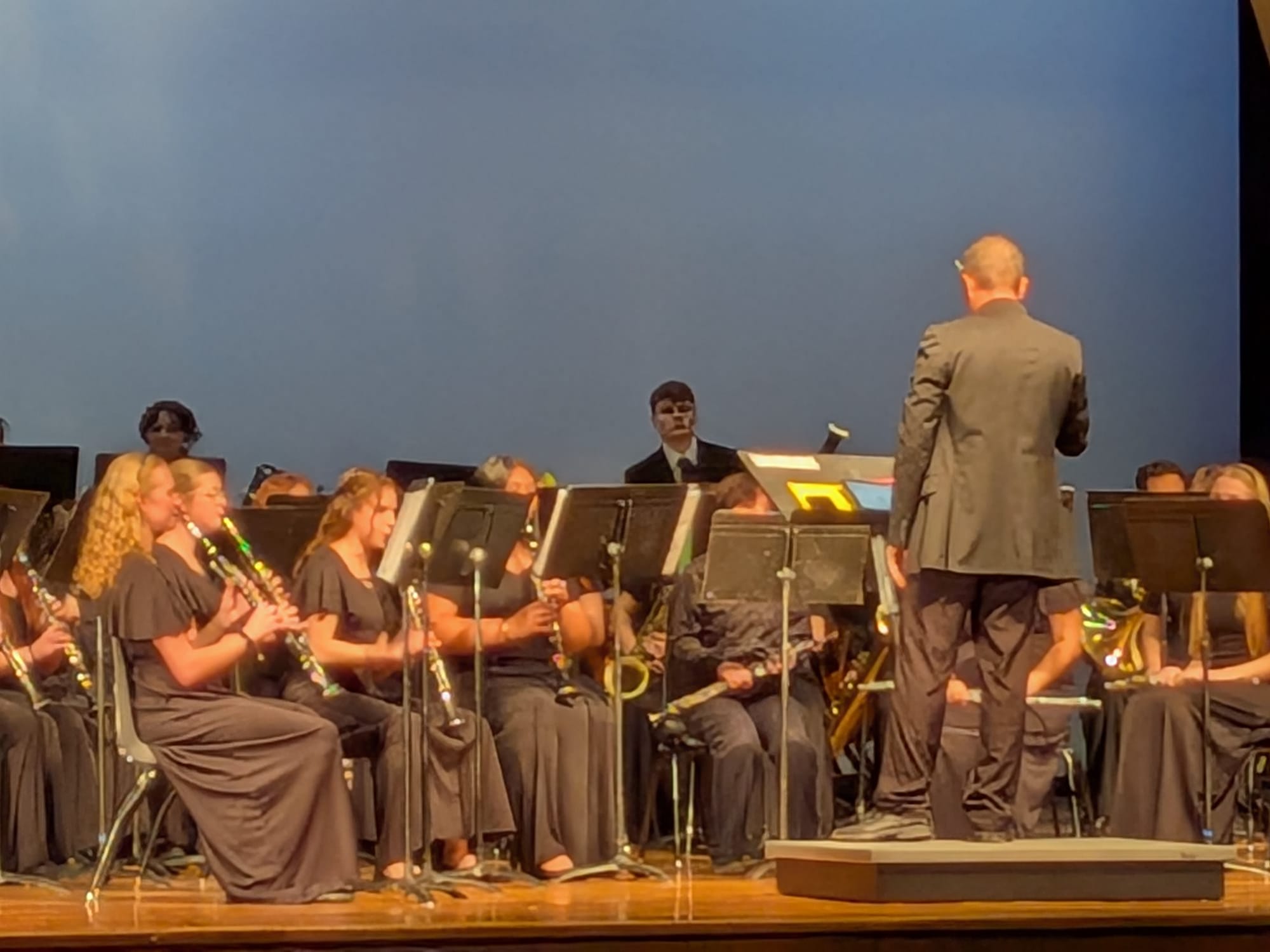 A 8x zoomed in photo of a concert band or orchestra performing on a stage. Musicians in black formal attire are seated with woodwind instruments, and a conductor in a grey suit is standing on a podium with his back to the camera.