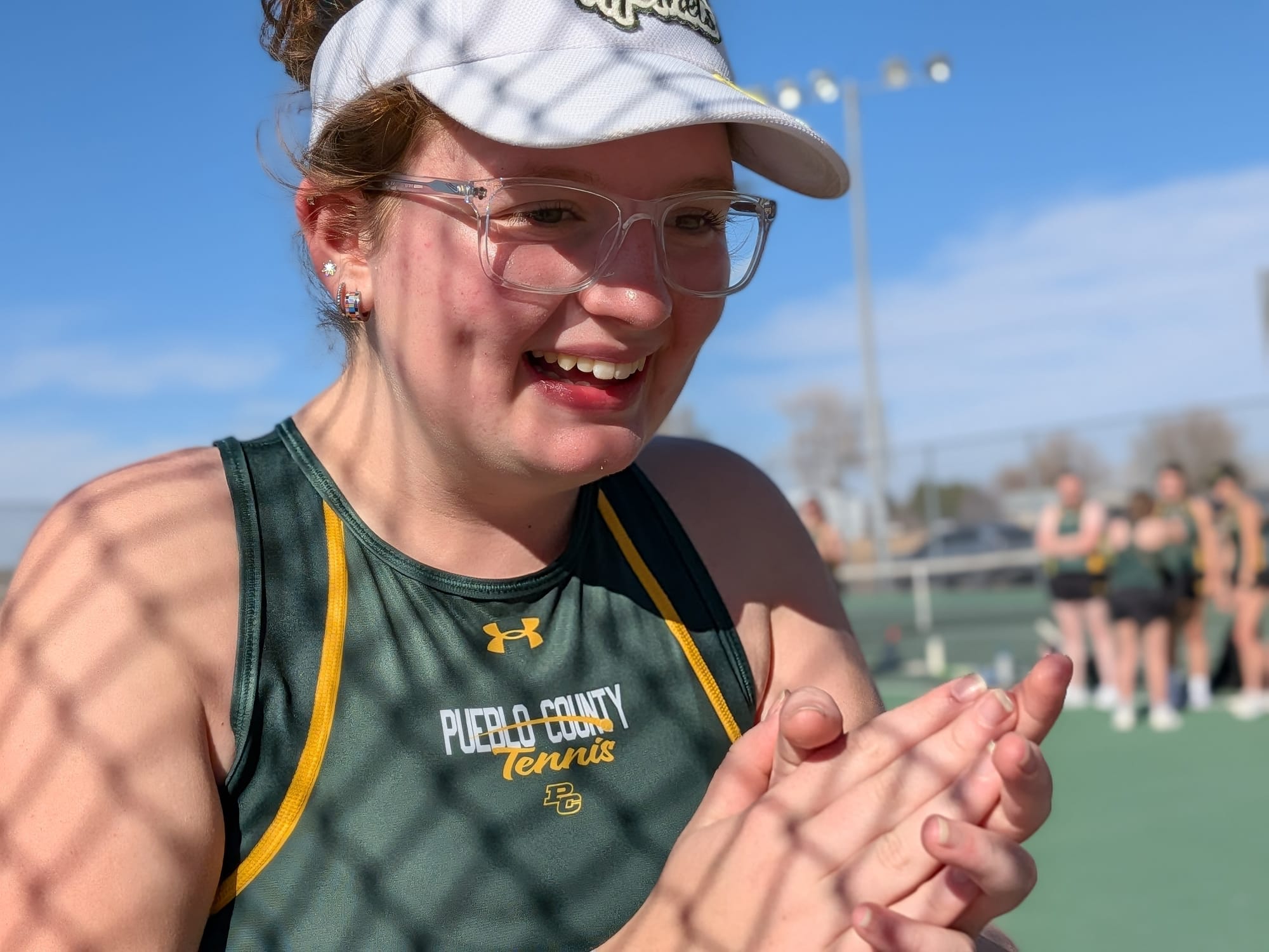 A portrait of a young woman smiling and clapping her hands. The photo is taken through a chain-link fence on a sunny day at a tennis court.