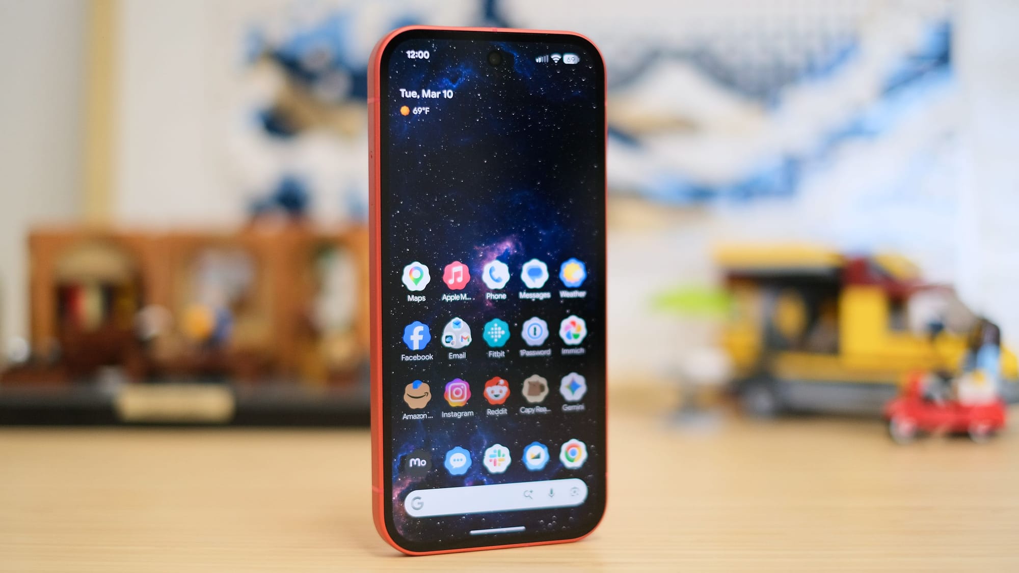 A front-facing view of a vibrant coral-colored smartphone standing upright on a wooden surface. The screen displays a home screen with various app icons over a dark, starry nebula wallpaper. The background is softly blurred, showing Lego sets.