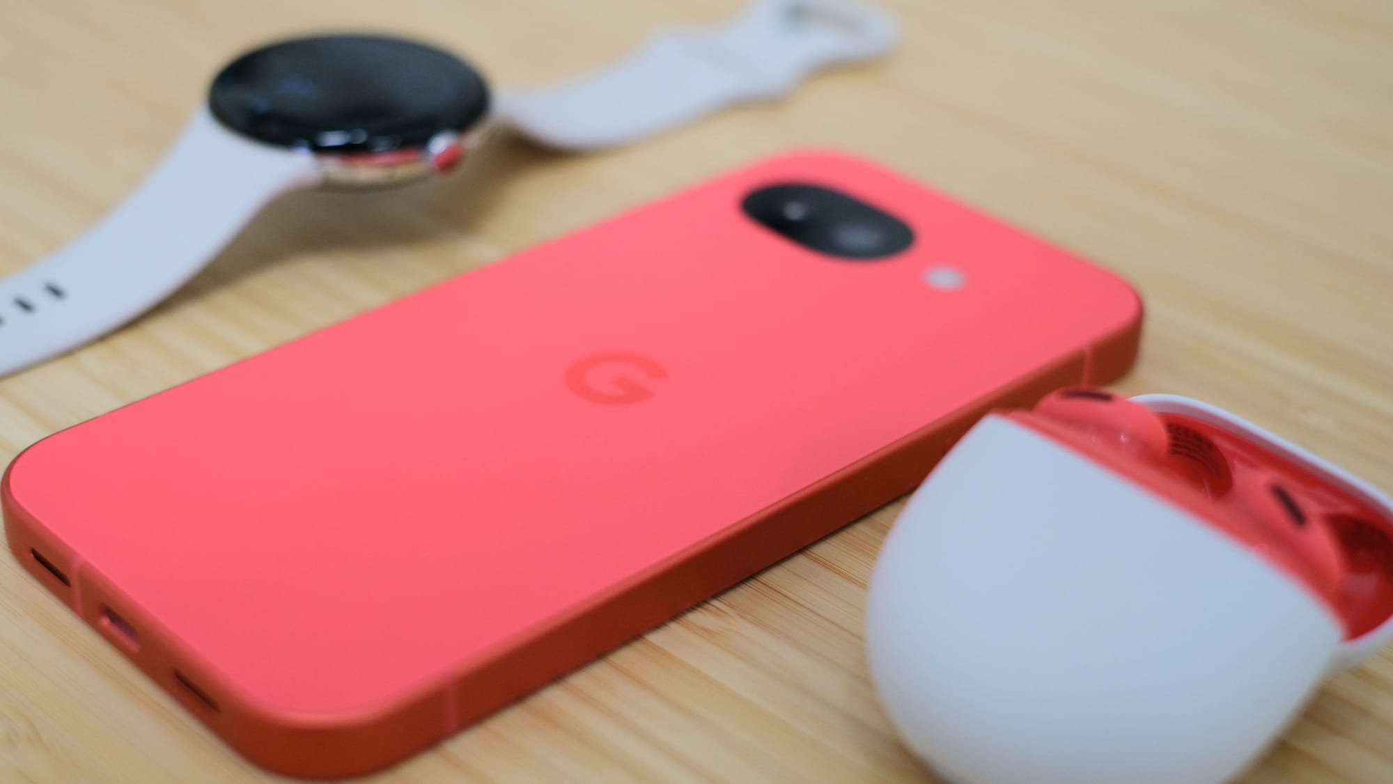 An angled tabletop shot showing the coral phone lying flat next to Pixel Watch 4 LTE and a pair of color-matching Pixel Buds 2a.