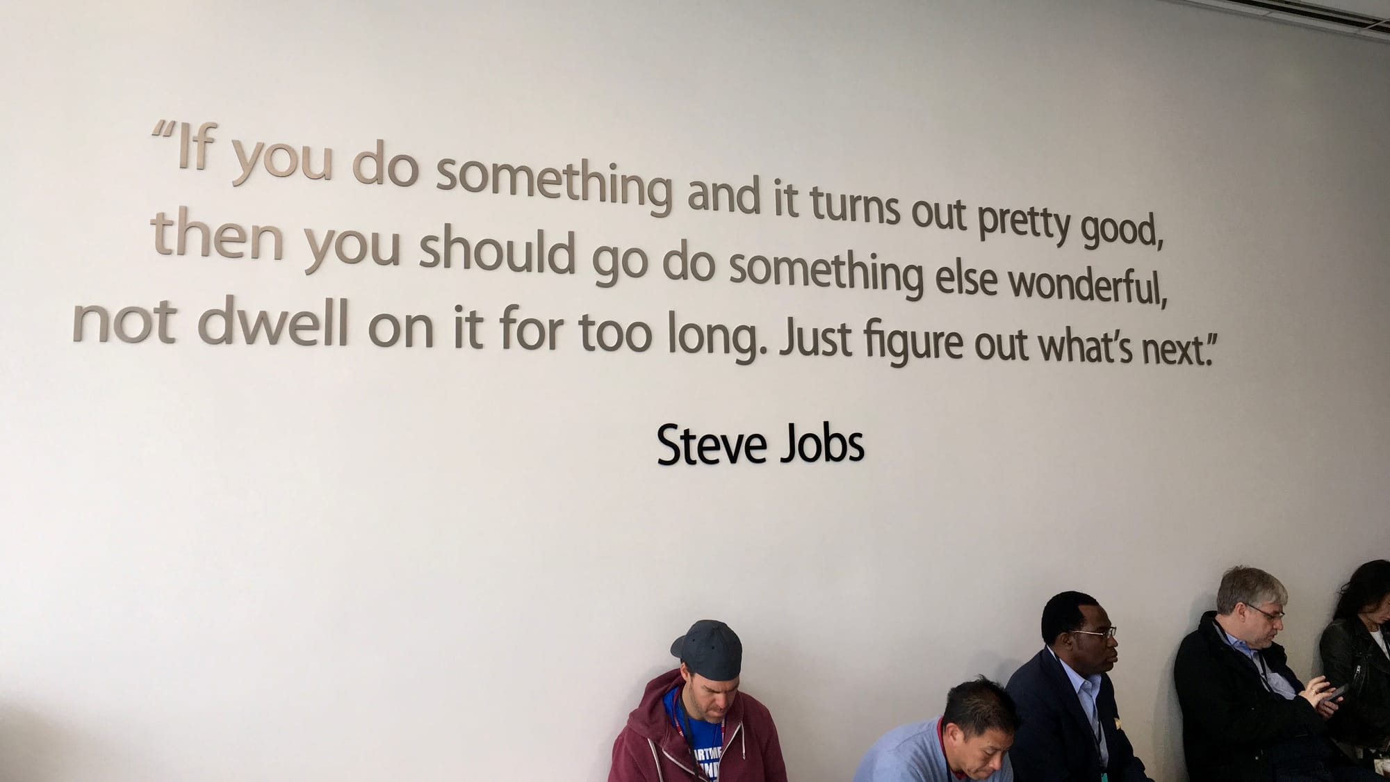 Steve Jobs quote on the wall at Apple's Town Hall about figuring out what's next, with attendees sitting below on their phones.